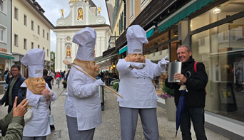 Drei Künstler von Rico Art Entertainment in übergroßen Comedy-Koch-Kostümen interagieren humorvoll mit einem lachenden Passanten auf einer Einkaufsstraße.