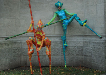 Zebra Stelzentheater Künstler in fantasievollen, blauen und orangefarbenen Chamäleon-Kostümen posieren auf Stelzen an einer Steinwand.