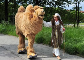 Walking Act von Rico Art Entertainment: Eine Dame in orientalischem Kostüm spaziert mit einem täuschend echten, lebensgroßen Kamel-Kostüm durch eine Parkanlage.